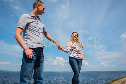 Пара в любви - стояли вместе, держась руками, рядом с водой в солнечный день, голубое небо с длинными белыми облаками на заднем плане. мужчины и женщины смотрят друг на друга, улыбаясь.
