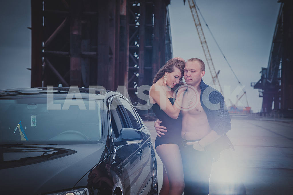 Beautiful blonde girl standing sexy on the unfinished bridge, with the light hair men and car, in black corset on a windy day, grey clouds, holding each other — Image 38990