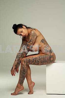 Young beautiful girl with tattoo posing nude in studio sitting on a white cube