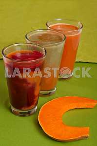 Various freshly squeezed vegetable juices with the hunk of pumpkin on green background.