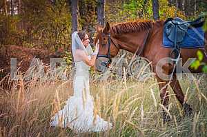 Brown-haired woman standing in front of a horse, in wedding dress mermaid silhouette, among the spikelet, autumn, forest on the background