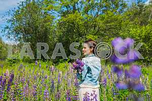 Young woman, brunette, happy, standing among the field of violet lupines, smiling, purple flowers. Blue sky on the background. Summer, sunny day! turned her back.