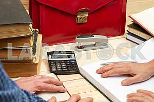 On a wooden table books, documents, calculator, red briefcase.