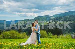 Wedding in mountains, a couple in love, mountains background, standing surrounded dandelions, among the lawn with the green grass, rustic style, girl in long tulle dress, romantic landscape, hugging