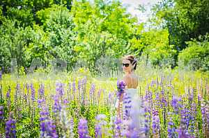 Young woman, happy, standing among the field of violet lupines, smiling, purple flowers. Blue sky on the background. Summer, sunny day, tuning back with bouquet