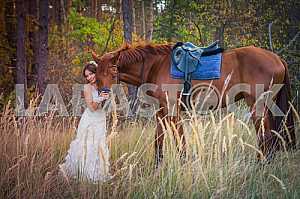 Brown-haired woman standing near the horse, in wedding dress mermaid silhouette, among the spikelet, autumn, forest on the background