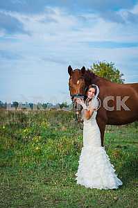 Brown-haired woman standing in front of a horse, in wedding dress mermaid silhouette, blue sky on the background