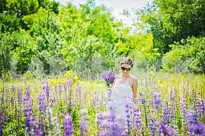 Young woman, happy, standing among the field of violet lupines, smiling, purple flowers. Blue sky on the background. Summer, sunny day! with bouquet