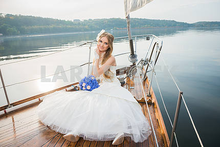 beautiful blonde bride in a long white dress poses on a sailing yacht at sea with the blue bouquet in her hand and the other hand holding herself on a sunny day
