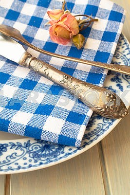 Silver fork and knife with an empty vintage plate on a blue napkin, with dry rose