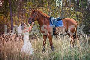 Bride brown-haired woman standing in front of a horse, in wedding dress mermaid silhouette, among the high autumn grass, autumn, forest on the background