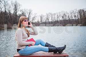 Young Woman speaking by the phone, with book on her hands, daily lifestyle, river on the background, spring, sunny day, sliming