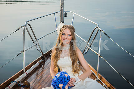 Beautiful blonde bride in a long white dress poses on a sailing yacht at sea with the blue bouquet in her hand, portrait, on a sunny day