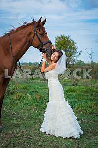 Brown-haired woman standing in front of a horse, in wedding dress mermaid silhouette, blue sky on the background
