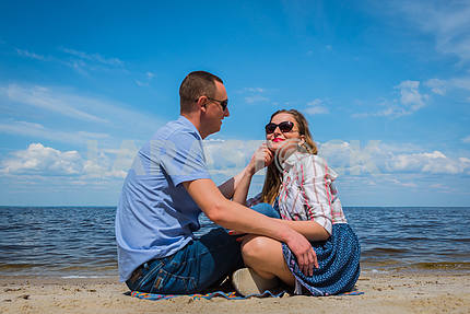 A loving couple sittingon the beach, near the water, playing with each other, blue sky on the background, sunny day, wide angle, long white clouds