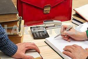 On a wooden table books, documents, calculator, red briefcase.