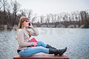 Young Woman speaking by the phone, with book on her hands, daily lifestyle, river on the background, spring, sunny day