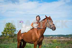 Bride brown-haired woman on a horse, in wedding dress, forest and blue sky on the background