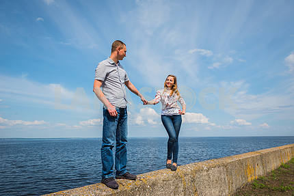 a couple in love - standing together, holding with the hands, near the water on a sunny day, blue sky with long white clouds on the background. men and women are looking into each other, smiling.