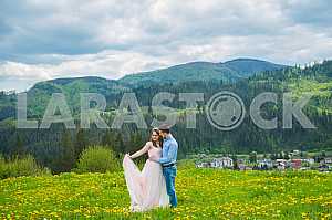 Wedding in mountains, a couple in love, mountains background, standing surrounded dandelions, among the lawn with the green grass, rustic style, girl in long tulle dress, romantic landscape
