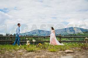 Newlyweds - handsome groom and gorgeous bride walking on trail - to each other, across field with forest hills and cloudly sky as background