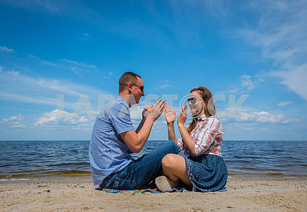 Влюбленная пара sittingon пляже, недалеко от воды, играя друг с другом, голубое небо на заднем плане, солнечный день, широкий угол, длинные белые облака