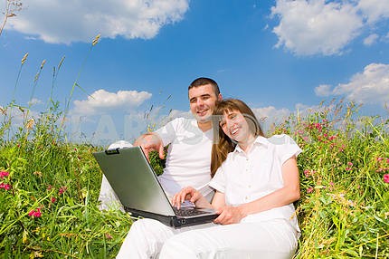 Young couple sitting on the grass field with a laptop. Against t