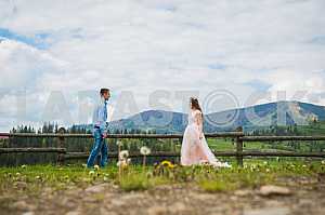 Newlyweds - handsome groom and gorgeous bride walking on trail across field with forest hills and cloudly sky as background, old wooden fence