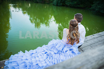 A love story couple, in love,  sitting back,  together in the forrest park, sitting  on the wooden bridge, girl in a beautiful violet dress, sunny evening, summer, green water on the background