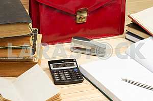 On a wooden table books, documents, calculator, red briefcase.
