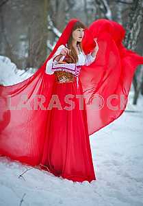 Women red ridding hood with blond hair wearing a long red cloak and old red dress walks in winter forrest, A lovely young girl, among trees and snow, holding a pies with basket, fairy tales