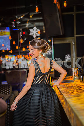 Beautiful brunette woman walking out in the restaurant, in black dress and red shoes. Smiling with her red lips, shy like a little girl showing her back