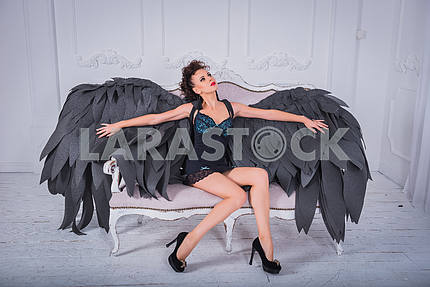 Brunette woman - black angel with the big wings, red lips, black short suit, in white room  sitting on the sofa