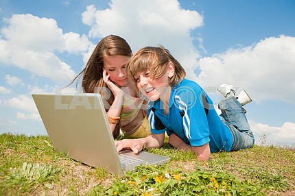 Two Smiling teenagers with laptop resting on meadow.