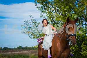 Brown-haired woman on a horse, in wedding dress, forest and blue sky on the background